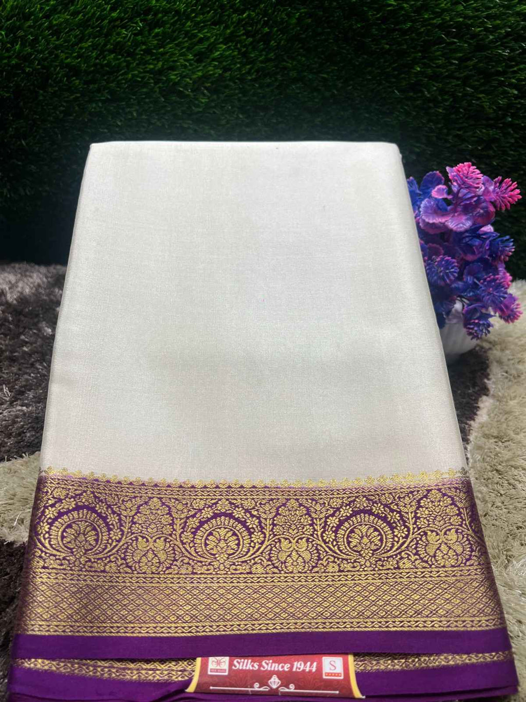 White saree with a purple and gold border on a textured surface with flowers in the background