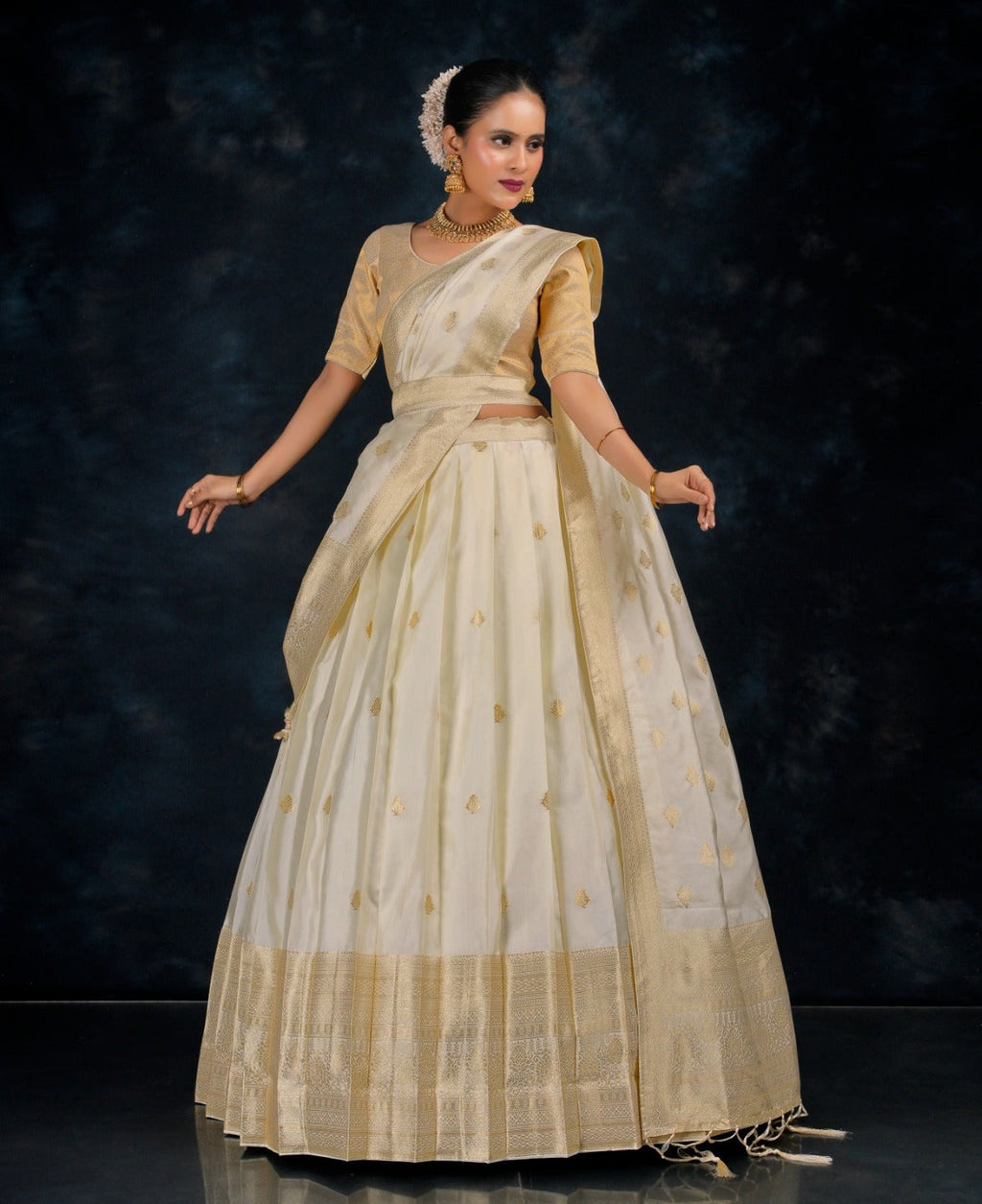 Woman wearing a traditional cream saree with gold border against a dark background