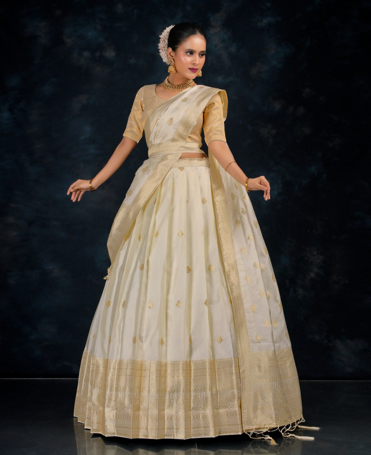 Woman wearing a traditional cream saree with gold border against a dark background